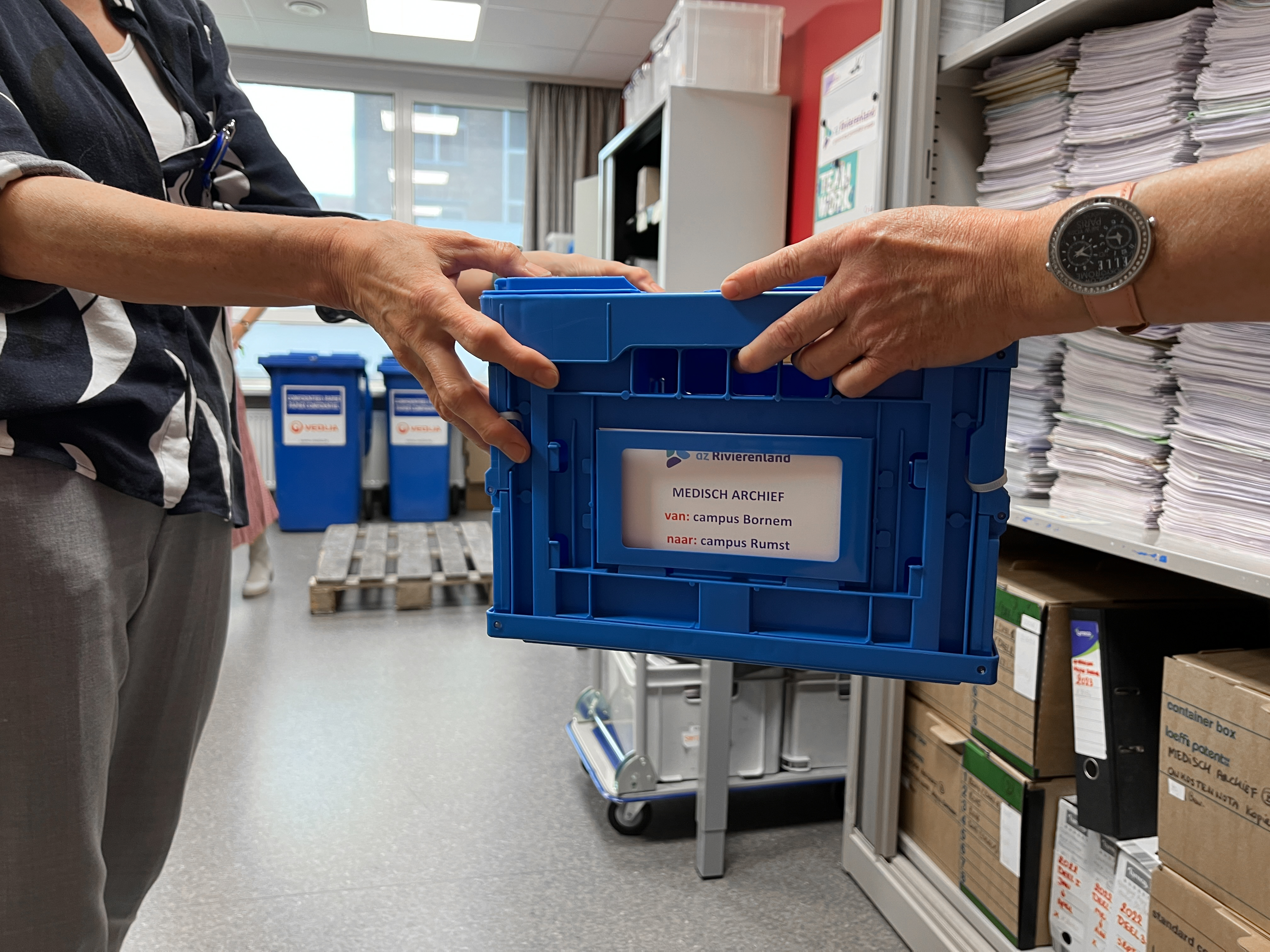 Een logistiek medewerker reikt een blauwe doos aan aan de collega's van het medisch archief.